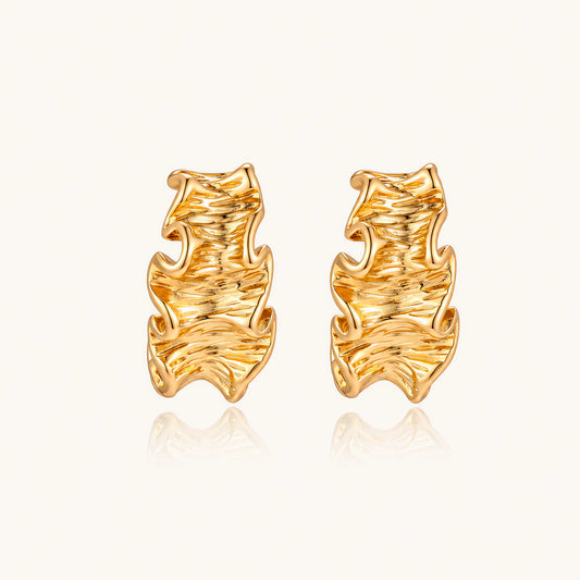 Pair of gold huggie hoops with sculpted wave texture and dimensional ripples on neutral background.