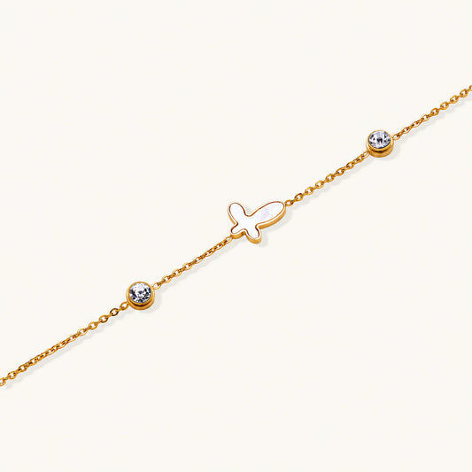 Gold chain bracelet with white mother of pearl butterfly pendant and two bezel-set crystals on cream background