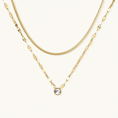 Layered gold necklace with herringbone and link chains, small bezel-set crystal pendant on cream background