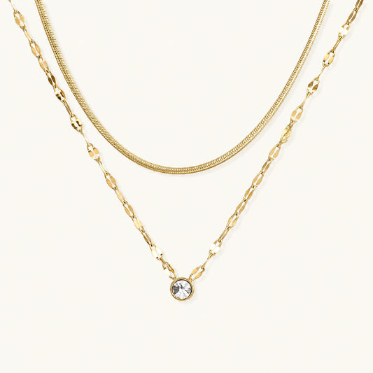 Layered gold necklace with herringbone and link chains, small bezel-set crystal pendant on cream background