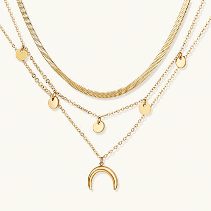Four-layer gold necklace with herringbone chain, disc charms, and crescent moon pendant on cream background