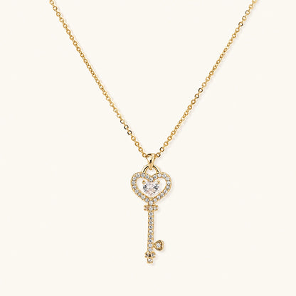 Delicate gold key pendant with heart-shaped bow and crystal pavé detailing on fine chain against cream background.