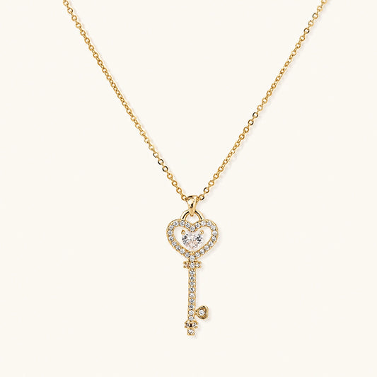 Delicate gold key pendant with heart-shaped bow and crystal pavé detailing on fine chain against cream background.