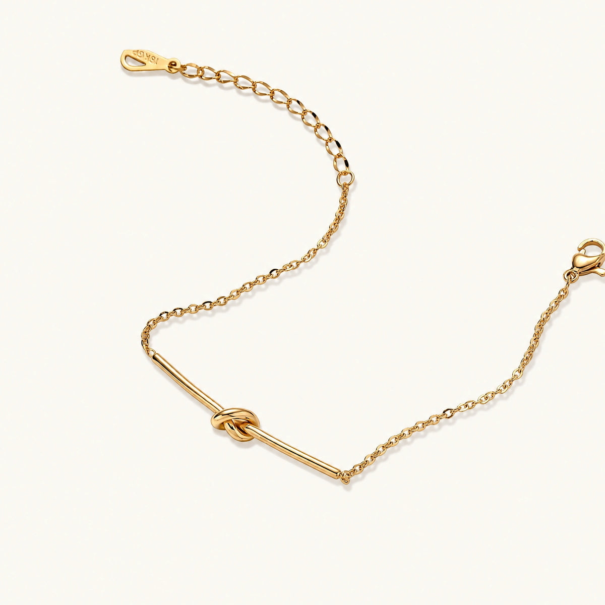 Delicate gold chain bracelet with minimalist bar and centered knot detail on cream background