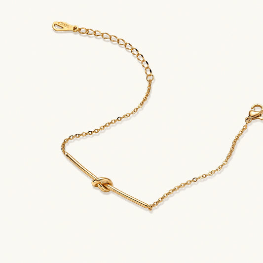 Delicate gold chain bracelet with minimalist bar and centered knot detail on cream background