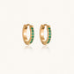 Pair of small gold huggie hoop earrings with bright green cubic zirconia stones on cream background