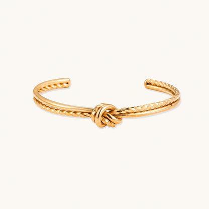 Gold cuff bracelet with twisted rope texture and decorative knot centerpiece on cream background.