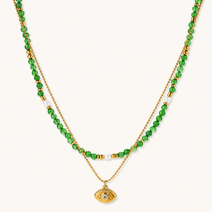 Layered necklace with green stone beads, gold spacers, white accents, and protective eye pendant on cream background.