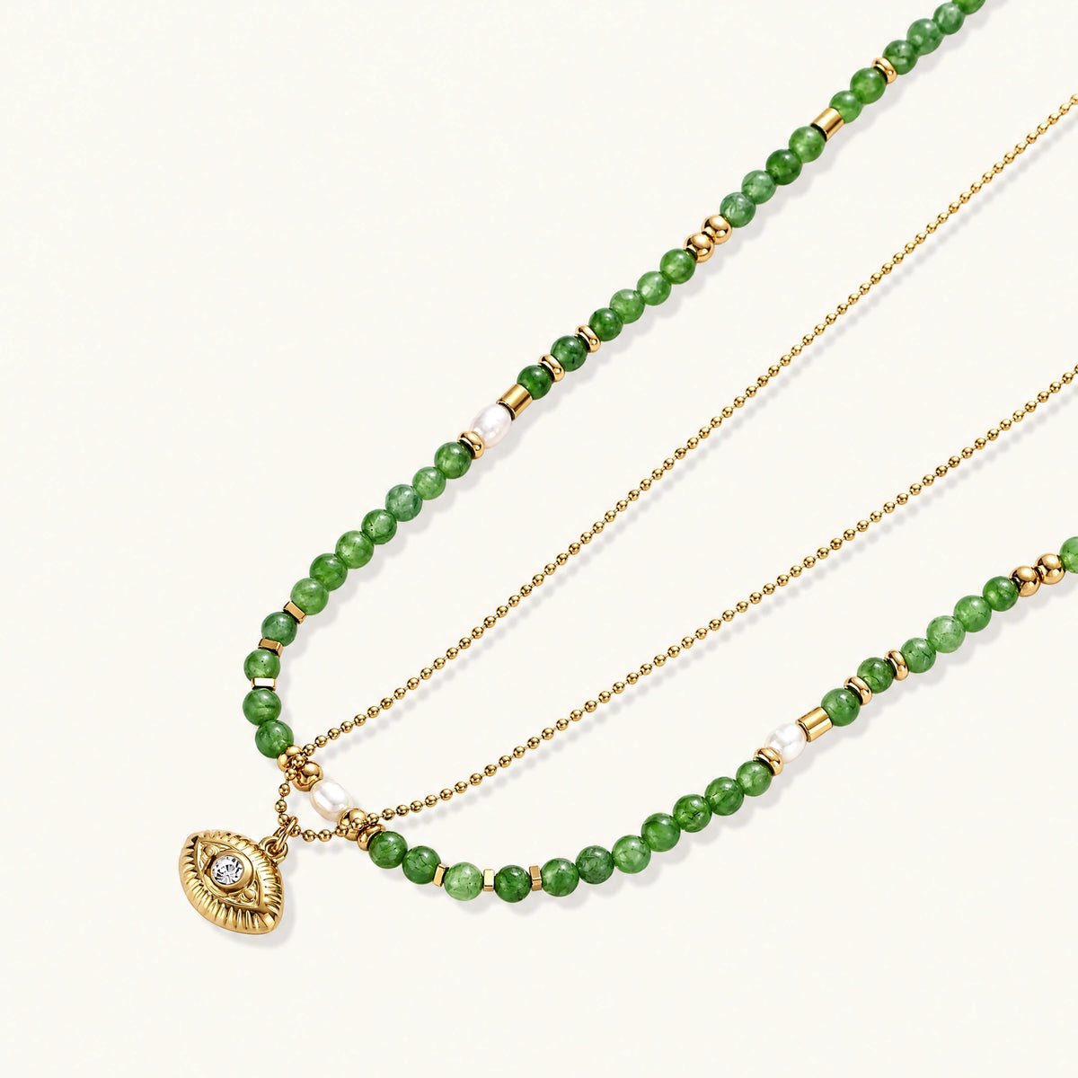 Layered necklace with green stone beads, gold spacers, white accents, and protective eye pendant on cream background.