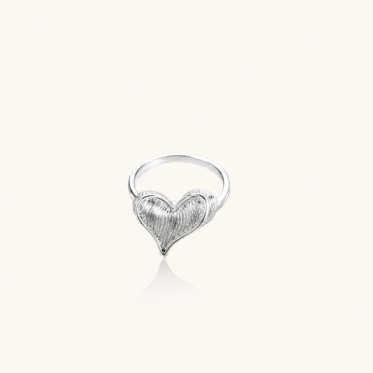 Silver ring with 3D heart formed by textured feather-like elements, white gold plated eco-brass on neutral background.