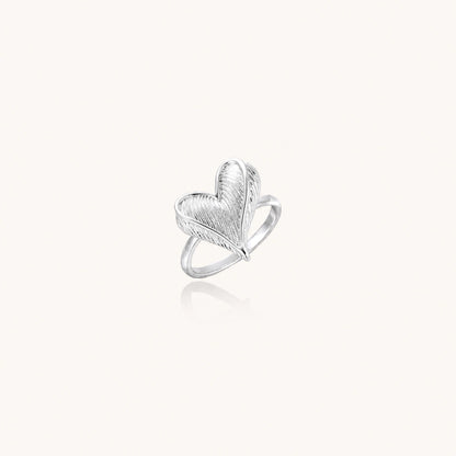 Silver ring with 3D heart formed by textured feather-like elements, white gold plated eco-brass on neutral background.