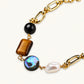 Gold chain bracelet with tiger's eye, obsidian, pearl, and iridescent abalone stones on cream background.
